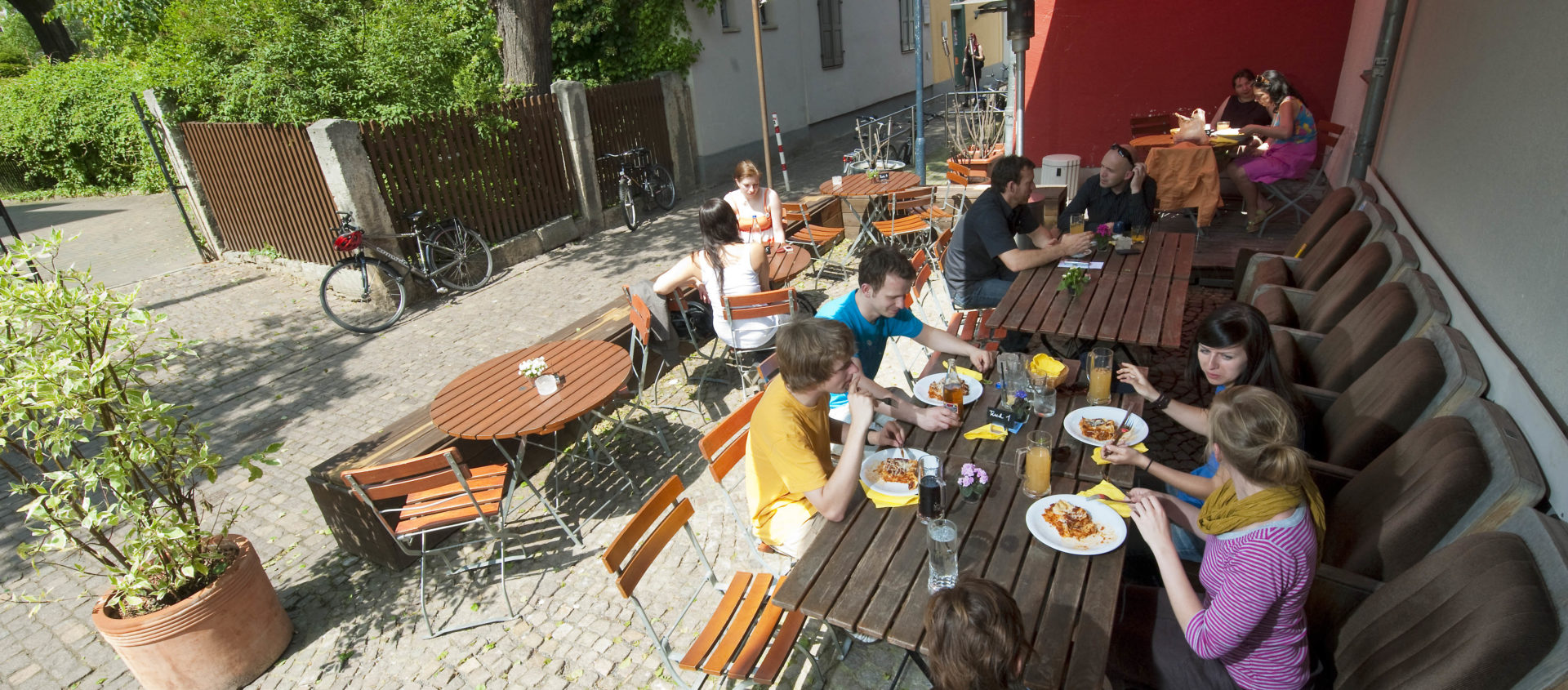 Mehrere Personen sitzen an langen Holztischen mit Stühlen und gepolsterten Bänken im Außenbereich des Theatercafés Jena und essen unter einem roten Sonnensegel auf gepflastertem Boden mit weiteren leeren Tischen und Stühlen in der Nähe