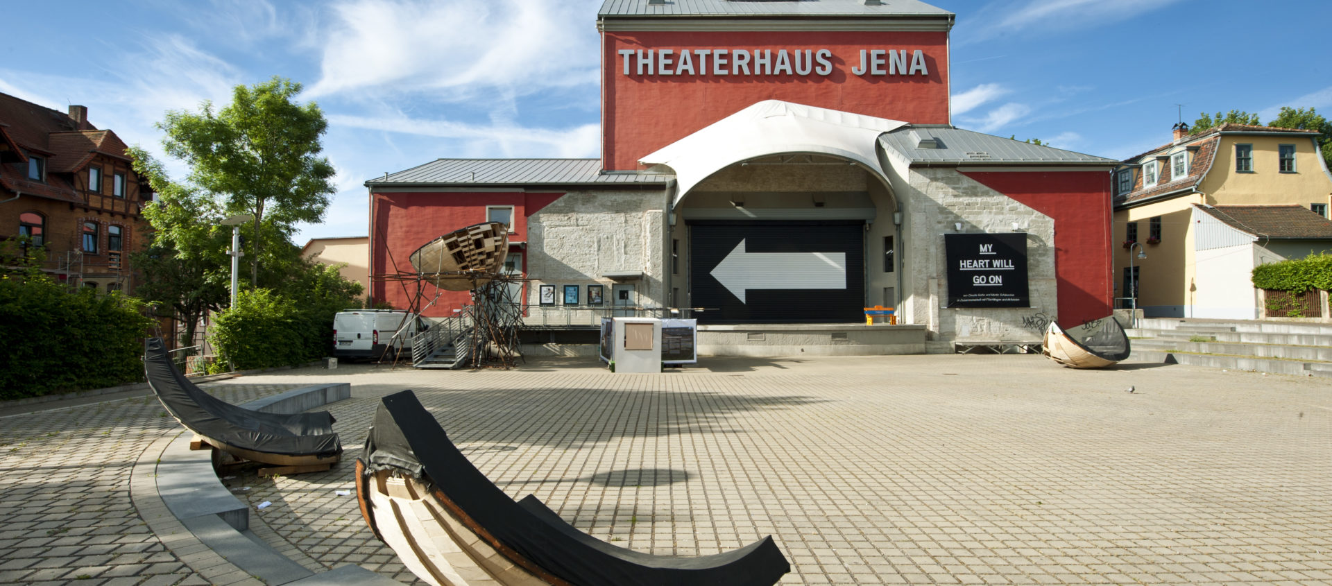 Das Theaterhaus Jena mit rotem Backsteingebäude, großem weißen Schriftzug und mehreren gebogenen Holzskulpturen auf dem gepflasterten Vorplatz