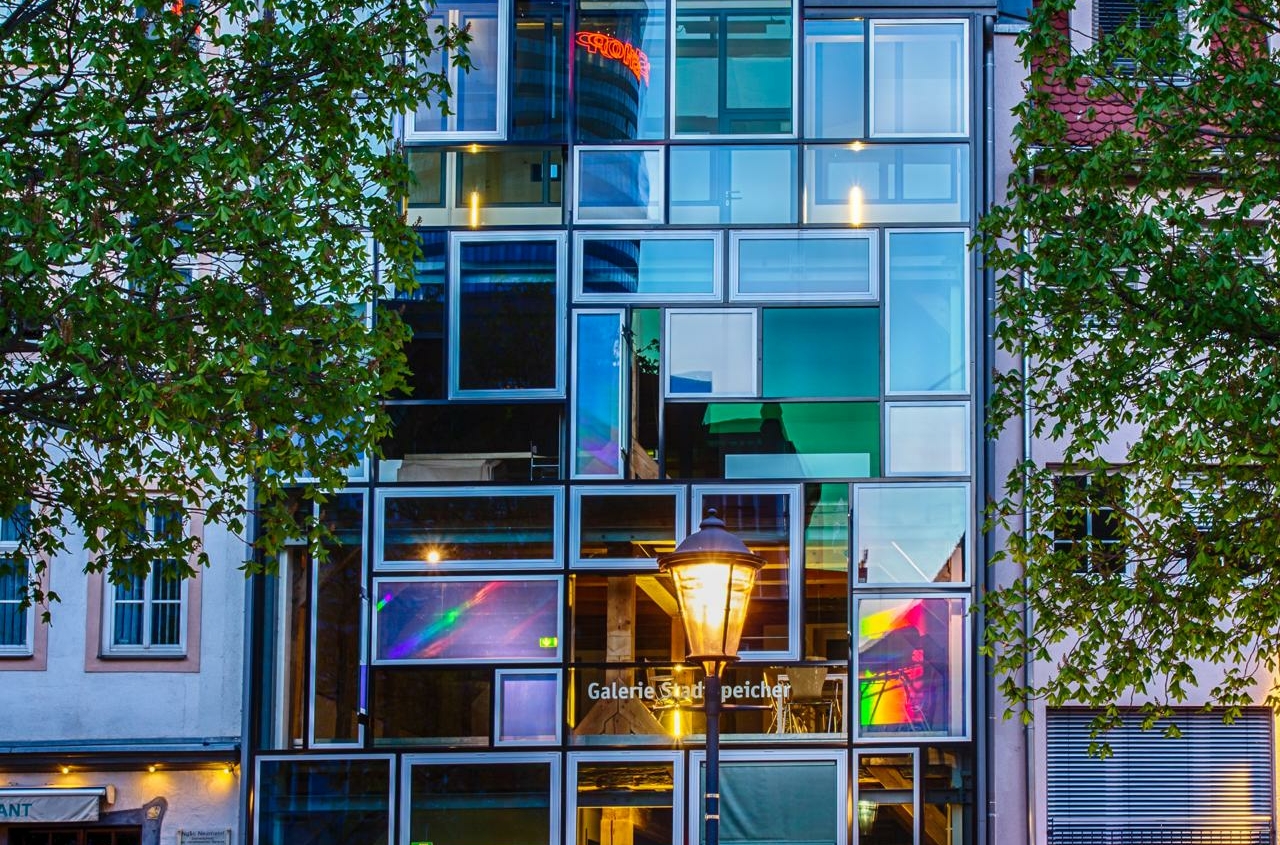 Fassade der Jena Tourist Information mit moderner Glasarchitektur und bunten Lichtreflexen, umgeben von historischen Gebäuden und einem Laternenpfahl im Vordergrund