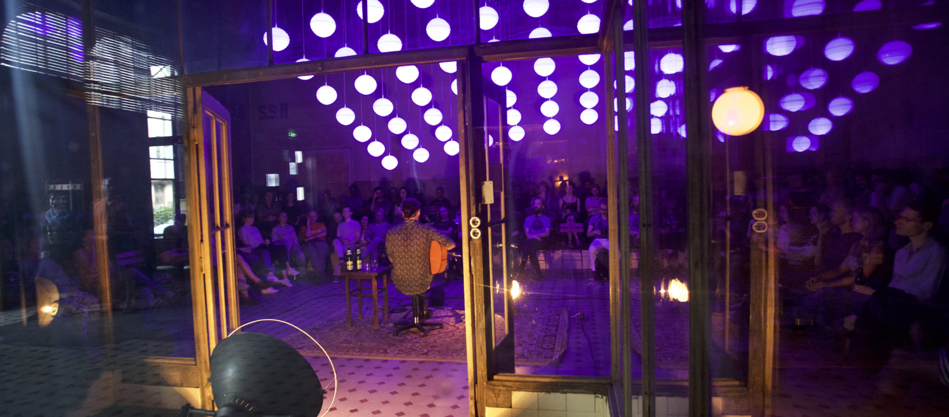 Bühne mit Musiker vor Publikum in einem Raum mit Decke aus vielen runden Lampen und Glaswänden