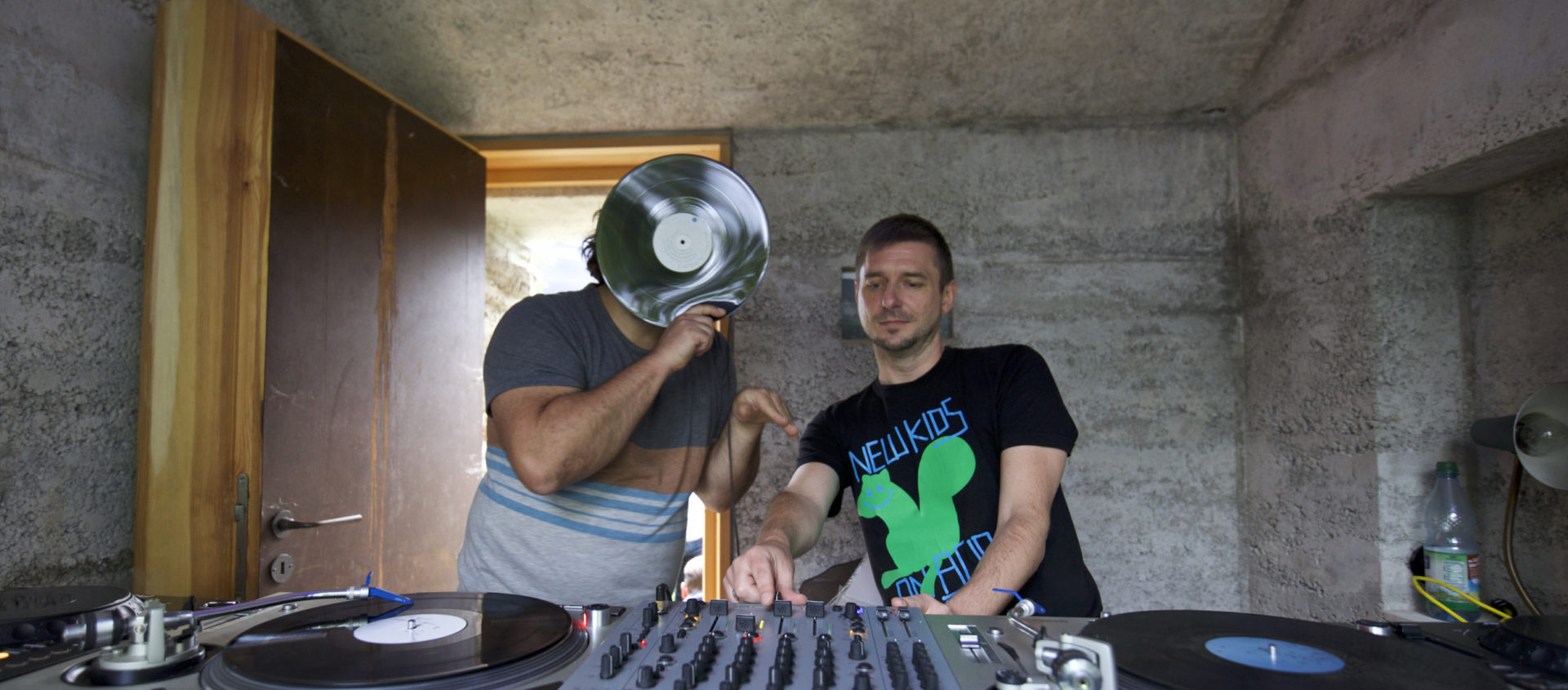 Zwei Männer stehen hinter DJ-Equipment in einem Raum mit rohen Betonwänden, einer hält eine Schallplatte vor sein Gesicht, der andere trägt ein T-Shirt mit der Aufschrift 'NEWKIDS'.