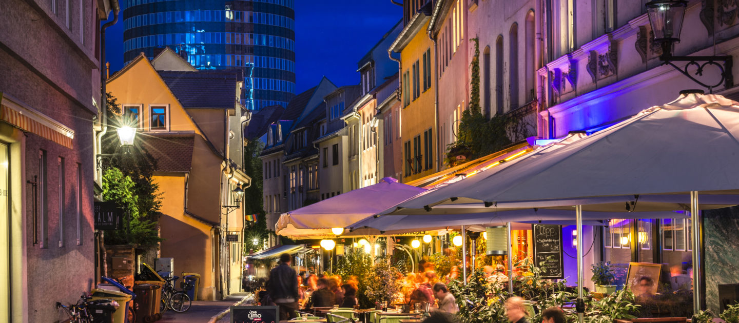Belebte Außengastronomie in der Wagnergasse in Jena mit Tischen, Sonnenschirmen und Menschen vor historischen Gebäuden bei Abendlicht