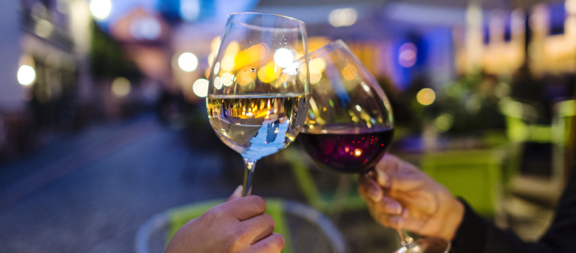 Zwei Hände stoßen mit einem Glas Weißwein und einem Glas Rotwein in einer abendlichen Straßenszene in Jena an