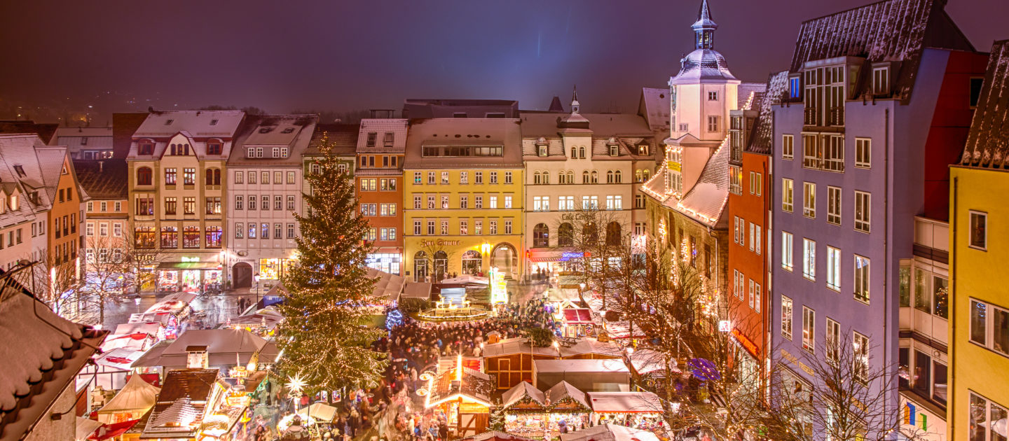 Luftaufnahme des Jenaer Weihnachtsmarkts mit festlich beleuchteten Ständen, großem Weihnachtsbaum und historischen Gebäuden im Hintergrund.