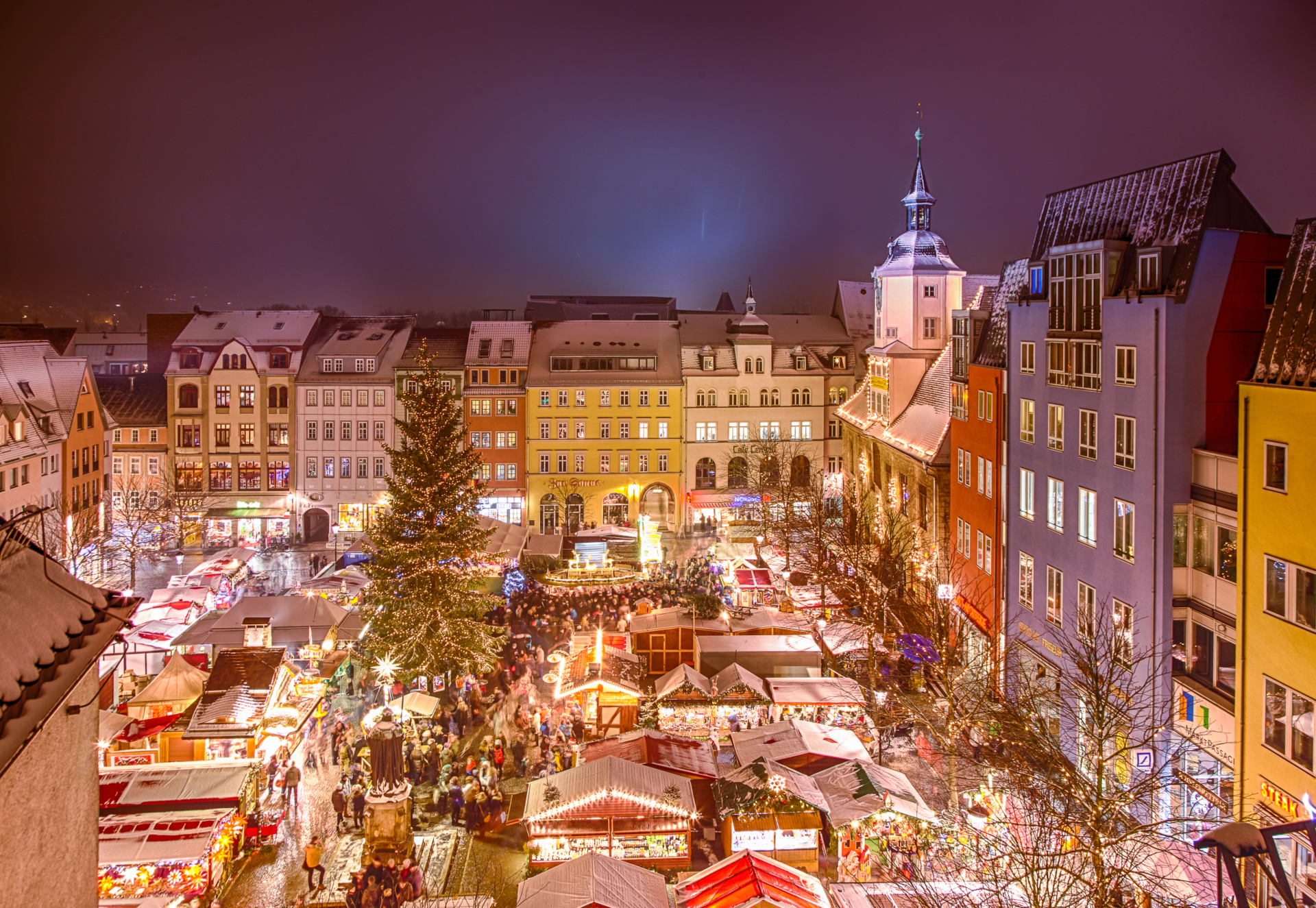 Luftaufnahme des Jenaer Weihnachtsmarkts mit festlich beleuchteten Ständen, großem Weihnachtsbaum und historischen Gebäuden im Hintergrund.