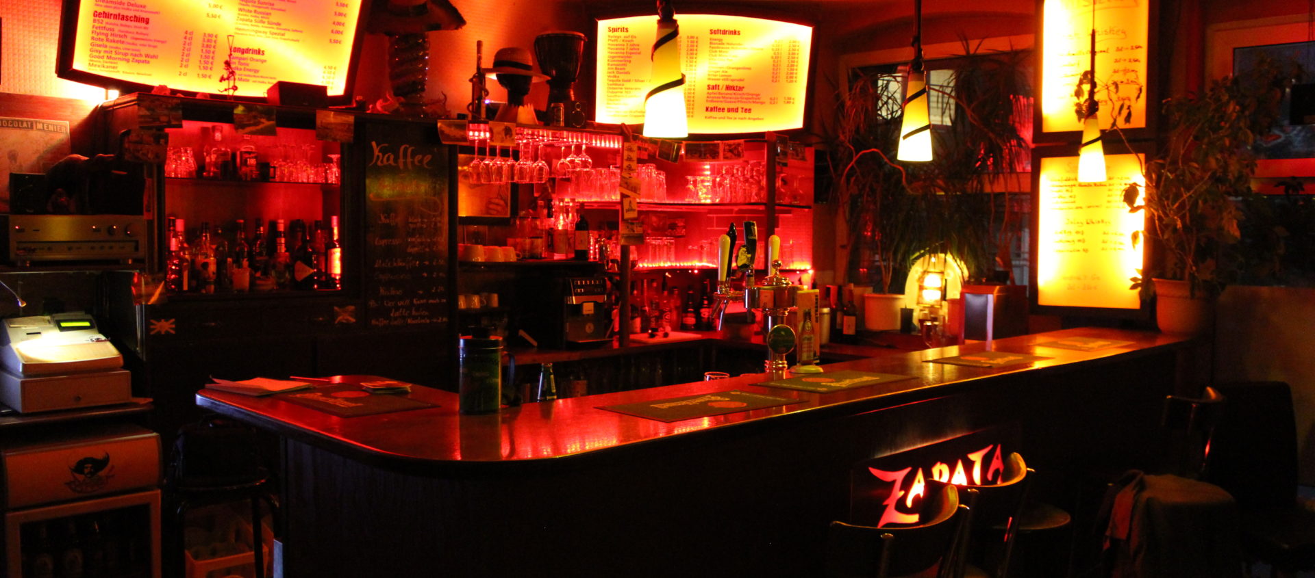 Dunkel belehrte Bar mit leuchtenden, gelblichen Menütafeln über dem Tresen und Stühlen mit der Aufschrift 'Zapata'