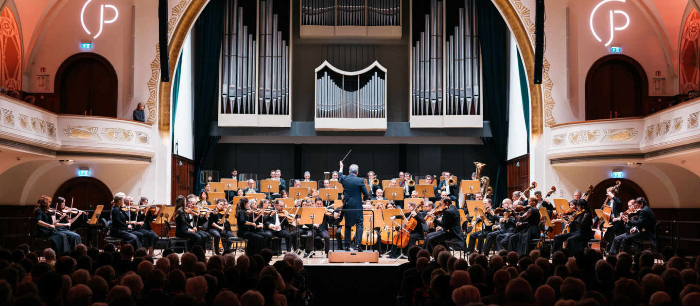 Orchester der Jenaer Philharmonie mit Dirigent auf Bühne in einem Konzertsaal mit großer Orgel im Hintergrund und Publikum im Vordergrund