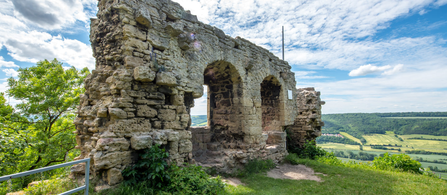 Ruine der Kunitzburg mit drei Rundbogenfenstern auf einem bewachsenen Hügel mit Blick auf ein Tal und bewaldete Hügel im Hintergrund