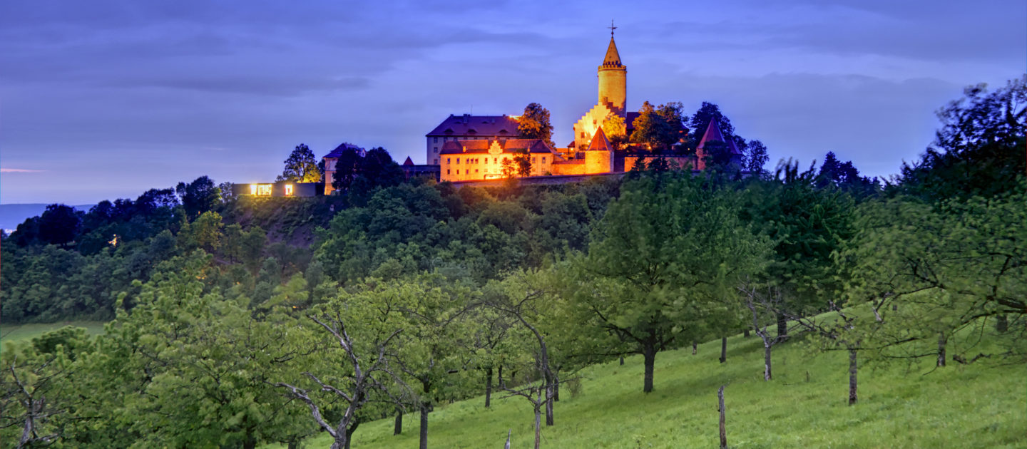 Die Leuchtenburg auf einem bewaldeten Hügel bei Dämmerung mit beleuchteten Gebäuden und einem Turm im Hintergrund und einer Wiese mit einzelnen Bäumen im Vordergrund