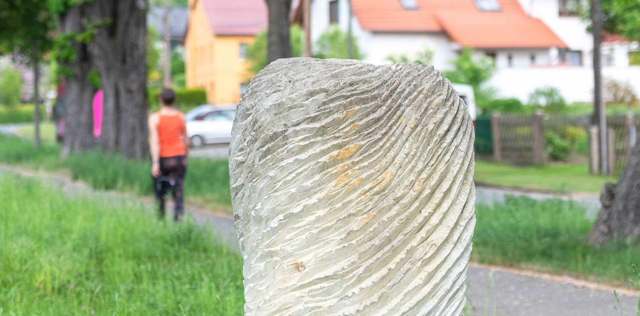 Skulptur mit wellenförmiger Struktur am Novalisweg in Jena, umgeben von Bäumen und einem Radfahrer im Hintergrund