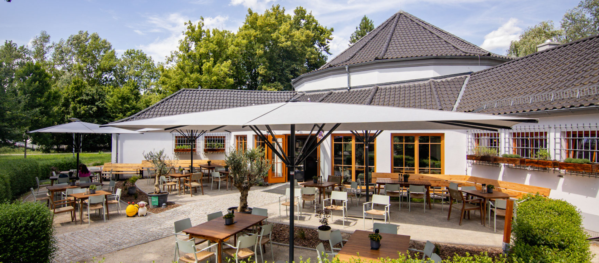 Außenbereich des Paradiescafés in Jena mit mehreren Holztischen und Stühlen unter großen Sonnenschirmen, umgeben von grünen Hecken und Bäumen