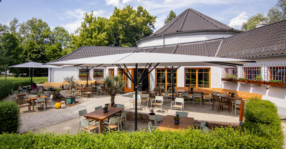 Paradiescafe-Jena-außen(3)-Bernd-Widemann Außenbereich des Paradiescafés in Jena mit mehreren Holztischen und Stühlen unter großen Sonnenschirmen, umgeben von grünen Hecken und Bäumen