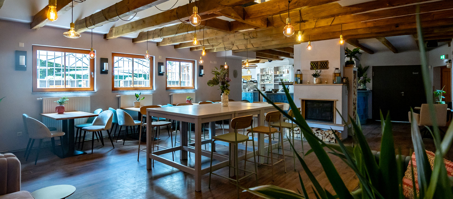Innenraum des Paradiescafés in Jena mit Holzbalkendecke, mehreren Tischen und Stühlen, Pflanzen und warmen Hängelampen.