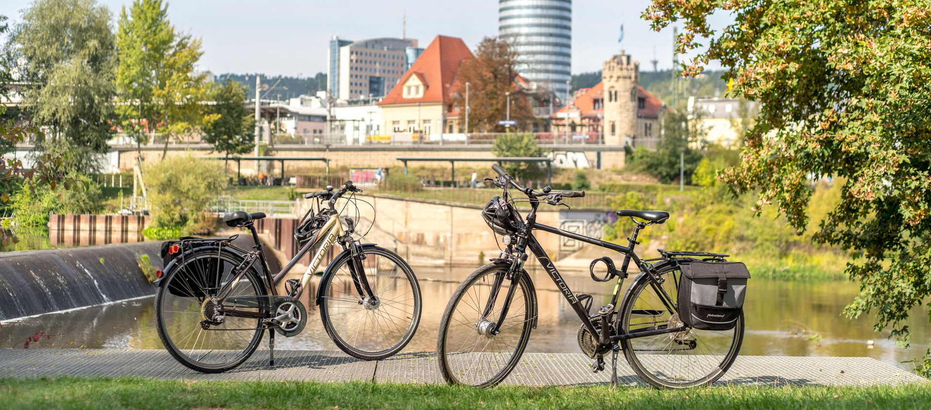 Zwei Fahrräder stehen im Paradiespark in Jena vor einem Wasserlauf mit Stadtgebäuden und dem JenTower im Hintergrund
