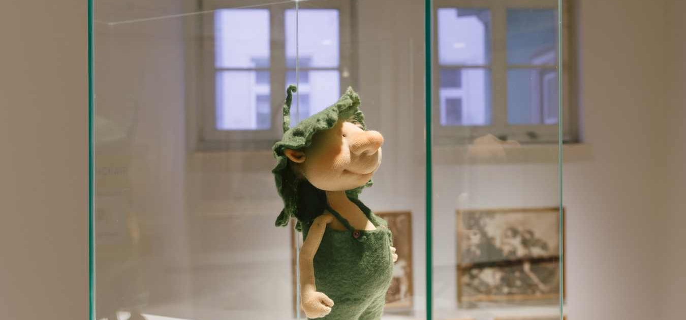 Grüne Stoffpuppe mit Zipfelmütze und Latzhose in einer Glaskassette im Romantikerhaus