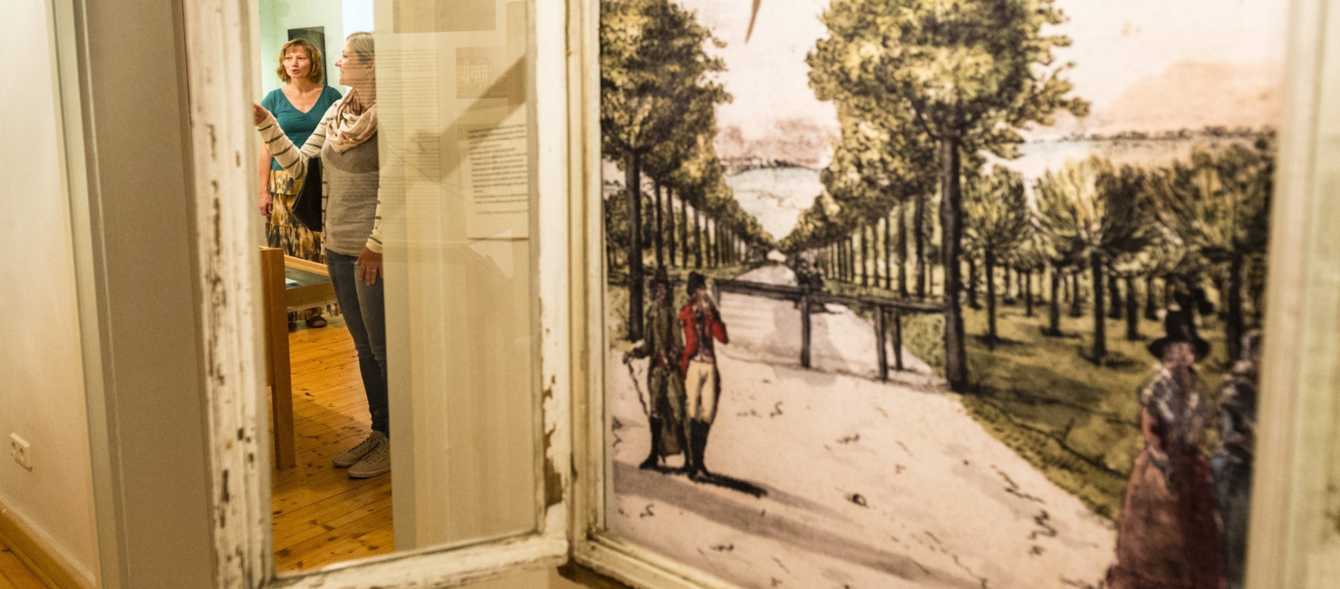 Blick durch einen offenen Fensterrahmen auf zwei Frauen in einem Raum des Romantikerhauses Jena, eine zeigt auf eine Wand mit Texten