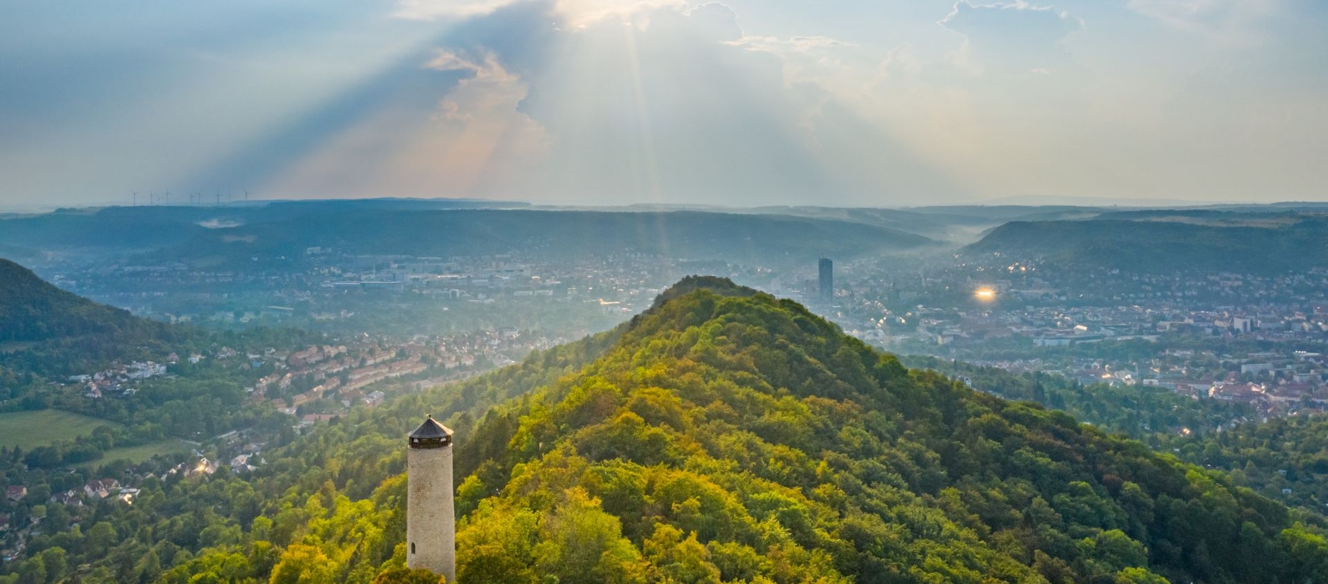 Blick auf den Fuchsturm auf einem bewaldeten Hügel mit der Stadt Jena im Hintergrund unter einem sonnigen Himmel