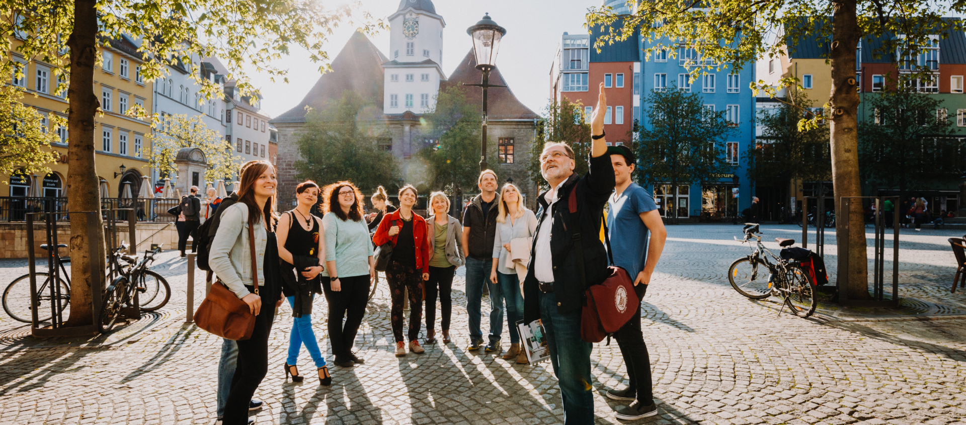 Gruppe von Menschen bei einer Stadtführung auf dem gepflasterten Marktplatz in Jena mit historischen Gebäuden und Bäumen im Hintergrund
