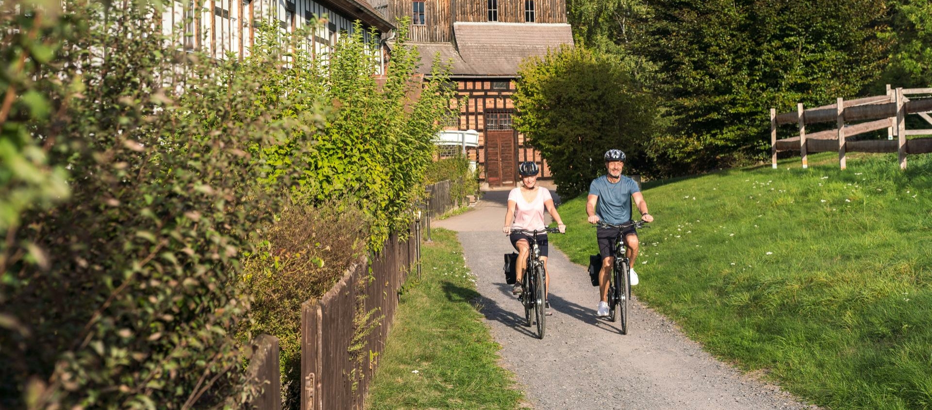 Zwei Radfahrer auf einem Weg neben einem Fachwerkhaus und einer Wiese mit Bäumen im Hintergrund