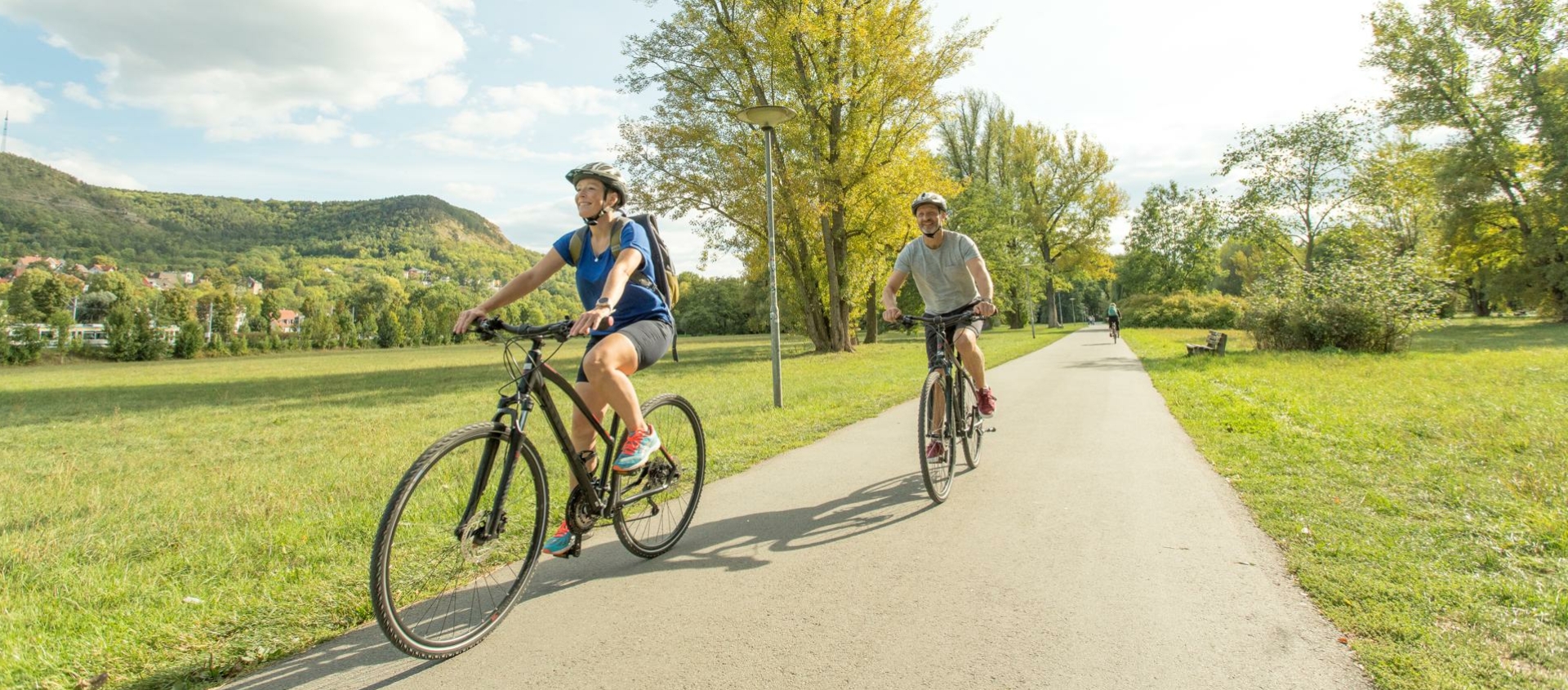 Zwei Radfahrer auf einem asphaltierten Weg im Paradiespark Jena, umgeben von Wiesen, Bäumen und Hügeln im Hintergrund.