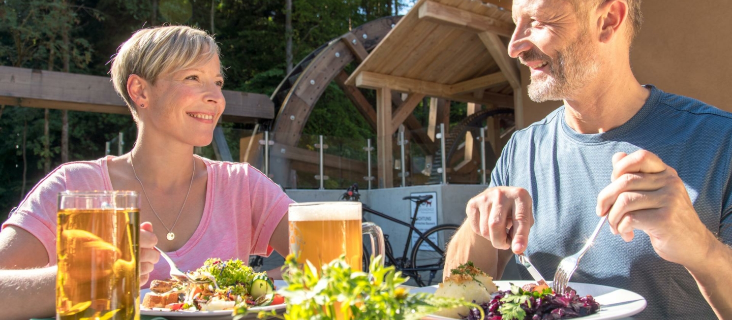Zwei Personen sitzen an einem Holztisch im Freien mit Biergläsern und Tellern mit Essen, im Hintergrund ein Wasserrad und Wald