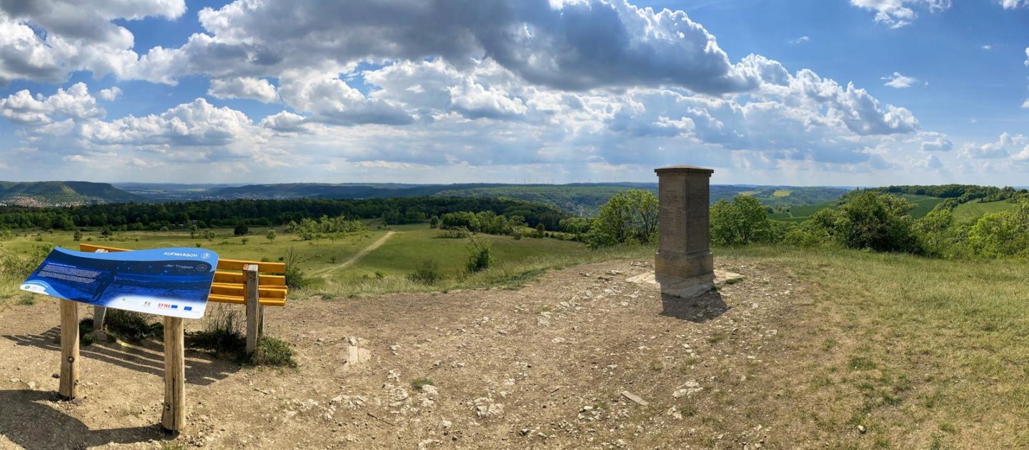 Napoleonstein auf einem Hügel bei Jena mit Infotafel und Bank, weiter Blick auf grüne Landschaft und bewölkten Himmel