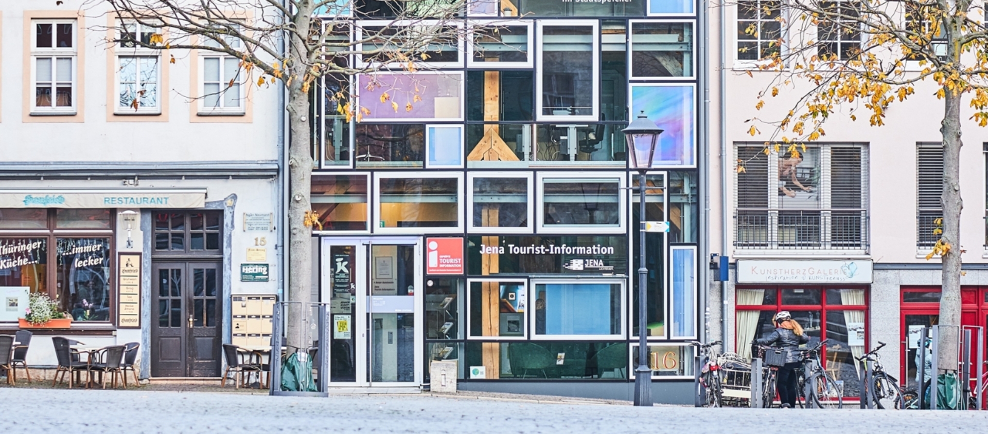 Moderne, schmale Fassade der Jenaer Tourist-Information mit großen Fenstern und umliegenden historischen Gebäuden am gepflasterten Marktplatz.