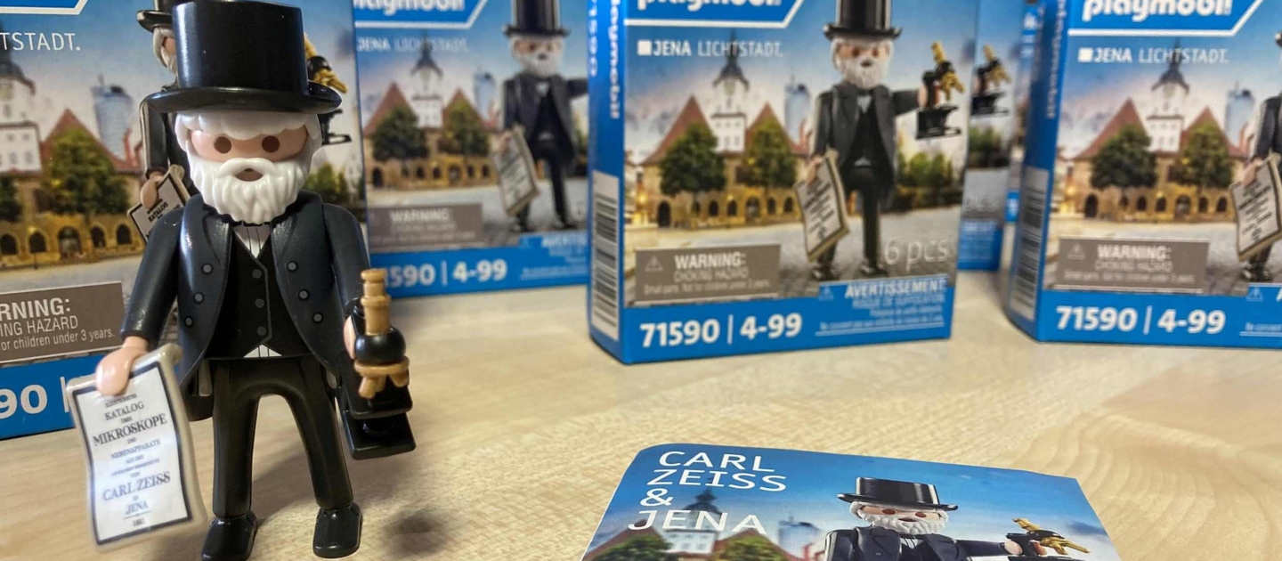 Playmobil-Sonderfigur von Carl Zeiss mit Zylinder, Bart, Mikroskop und Katalog, umgeben von Verpackungen und einem Faltblatt mit Aufschrift 'Carl Zeiss & Jena'