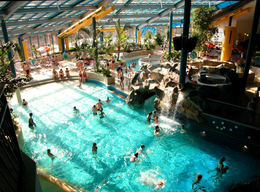 Innenansicht des Freizeitbades Galaxsea Jena mit mehreren Personen im Wasser und an den Beckenrändern, künstlichen Felsen und Wasserfall unter einem Glasdach