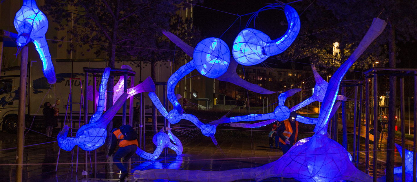 Leuchtende blaue, organisch geformte Skulpturen in einer nächtlichen städtischen Umgebung mit Bäumen und Gebäuden im Hintergrund