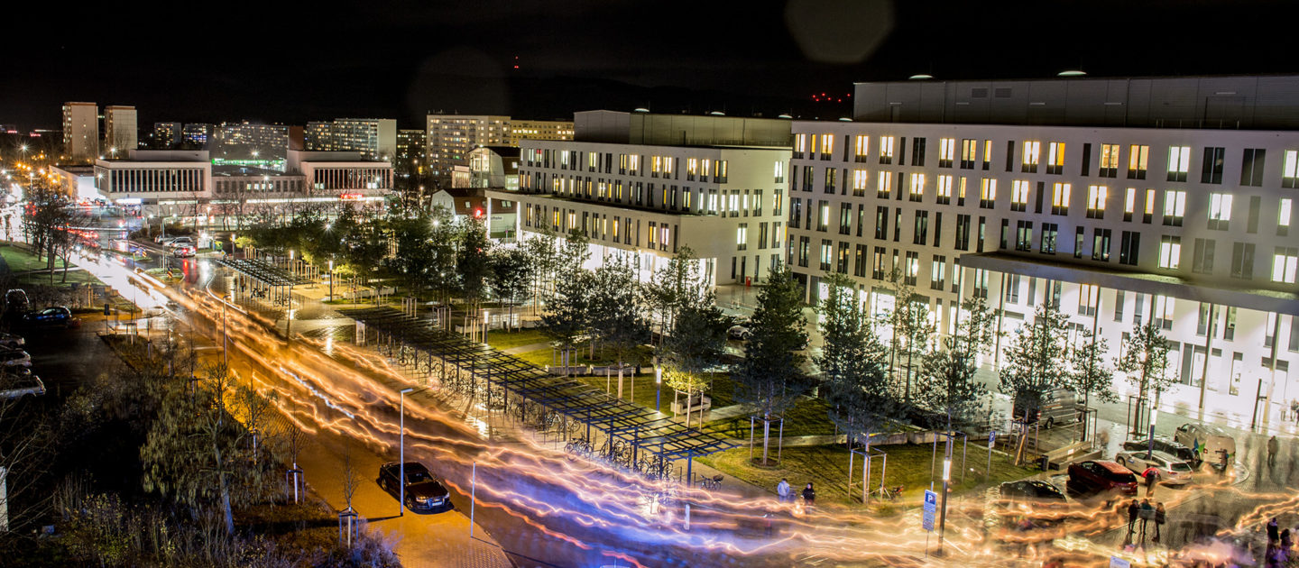 Nachtaufnahme einer belebten Straße in Jena mit beleuchteten Gebäuden, parkenden Autos und Lichtspuren von vorbeifahrenden Fahrzeugen