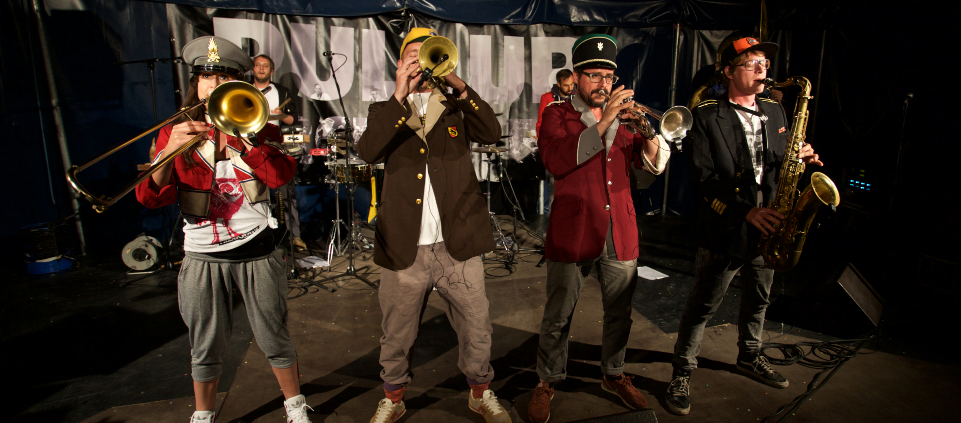 Vier Musiker der Wombo Orchestra spielen auf einer Bühne im Zirkuszelt bei der Jazzmeile, zwei mit Trompeten, einer mit Posaune und einer mit Saxofon, alle in bunter, teils legerer Kleidung.