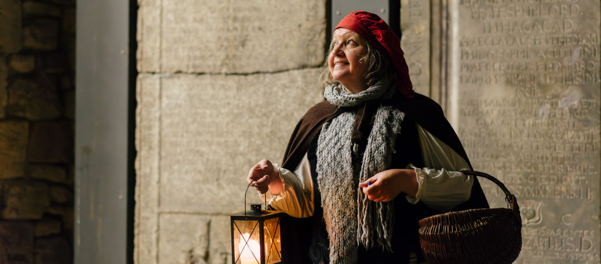 Frau in historischer Kleidung mit rotem Kopftuch, braunem Umhang und grauem Schal hält Laterne und Korb bei der Stadtführung 'Galgen, Gassen und Ganoven' in Jena