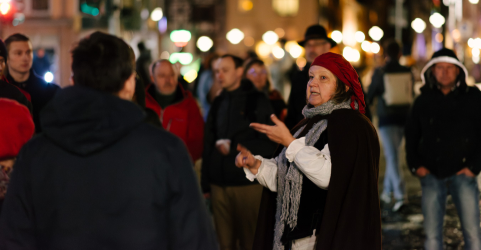 Frau in historischer Kleidung mit rotem Kopftuch führt abends Stadtführung 'Galgen, Gassen und Ganoven' in Jena vor einer Gruppe Zuhörer durch