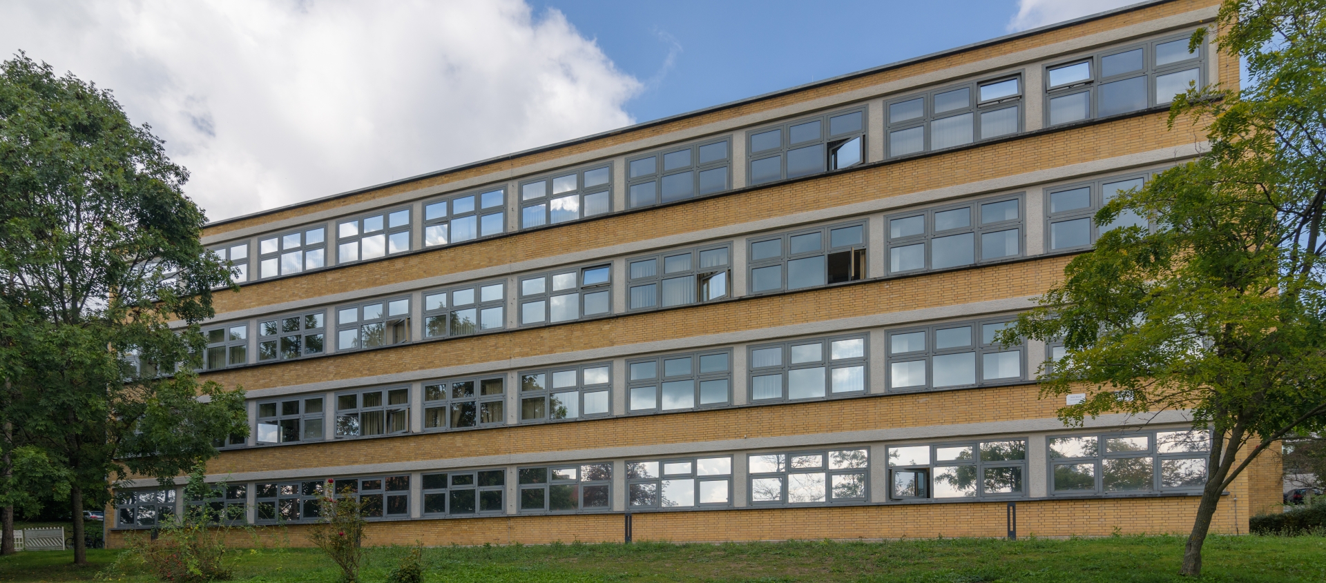 Mehrstöckiges, rechteckiges Gebäude mit vielen Fenstern und flachem Dach vor bewölktem Himmel und Rasenfläche mit Bäumen