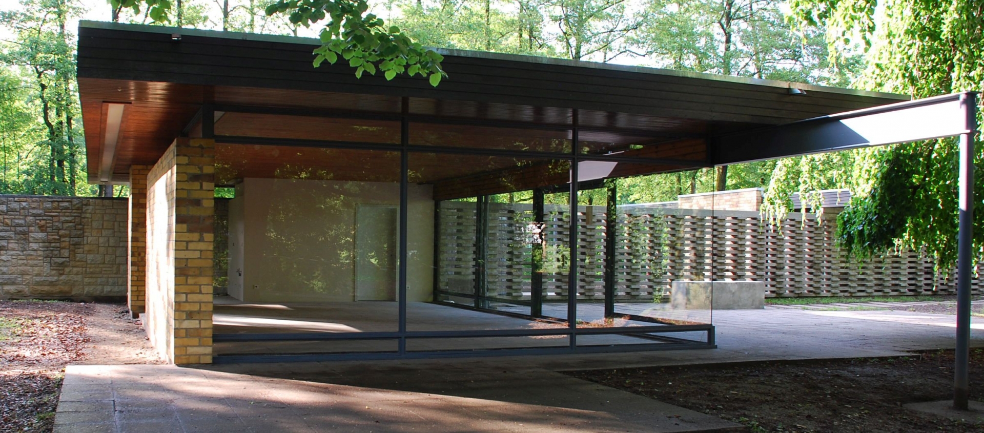 Modernes Glashaus mit Flachdach und großen Glasfronten, umgeben von Bäumen auf einer Rasenmühleninsel
