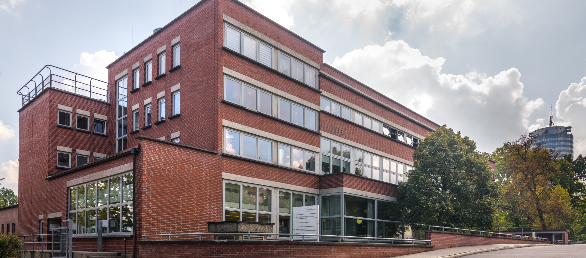 Mehrstöckiges Backsteingebäude mit großen Fenstern und flachem Dach vor bewölktem Himmel und Bäumen