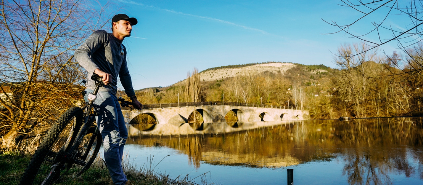 Person mit Fahrrad steht am Ufer der Saale nahe der Burgauer Brücke in Jena, im Hintergrund Hügel und klare Spiegelung der Brücke im Wasser
