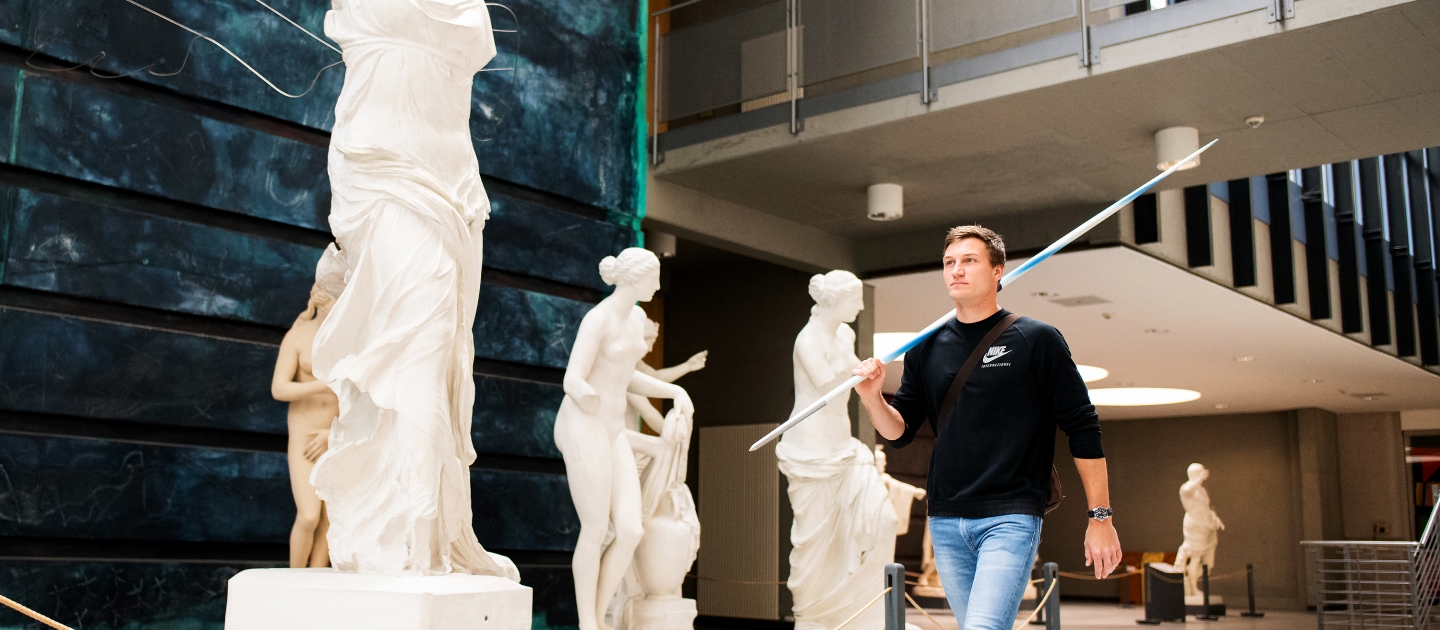 Person mit Langstock geht im Innenraum des Campus Jena an mehreren antiken Statuen vorbei