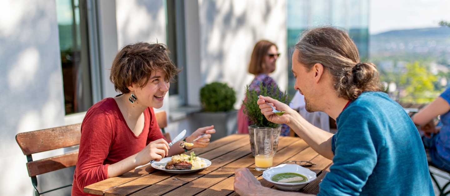 Zwei Personen sitzen an einem Holztisch auf einer Terrasse und essen, im Hintergrund weitere Personen und Glasfronten eines Gebäudes