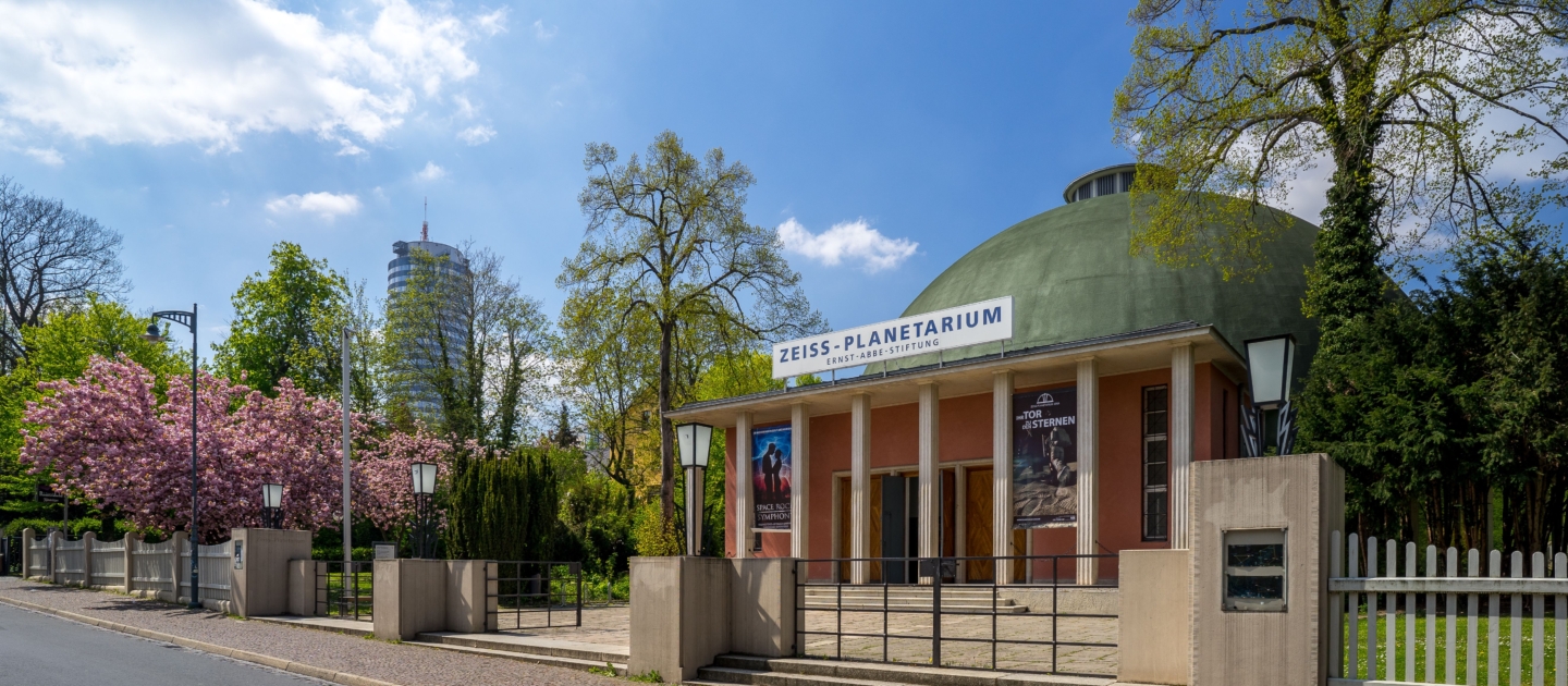 Zeiss-Planetarium mit grünem Kuppeldach und Säulen, umgeben von Bäumen und blühenden Sträuchern an einem sonnigen Tag