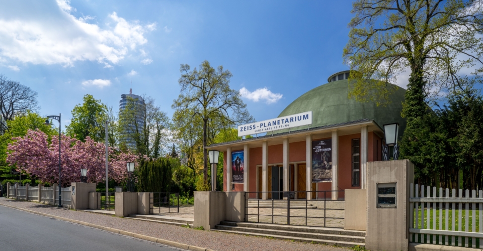 Zeiss-Planetarium mit grünem Kuppeldach und Säulen, umgeben von Bäumen und blühenden Sträuchern an einem sonnigen Tag