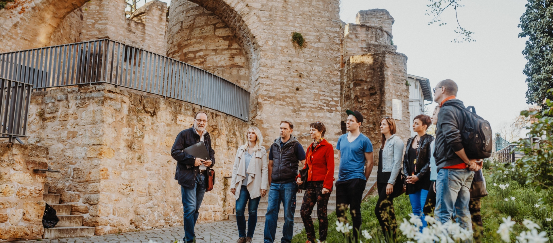 Gruppe von Menschen bei einer Stadtführung vor dem historischen Pulverturm in Jena mit blühenden Blumen im Vordergrund