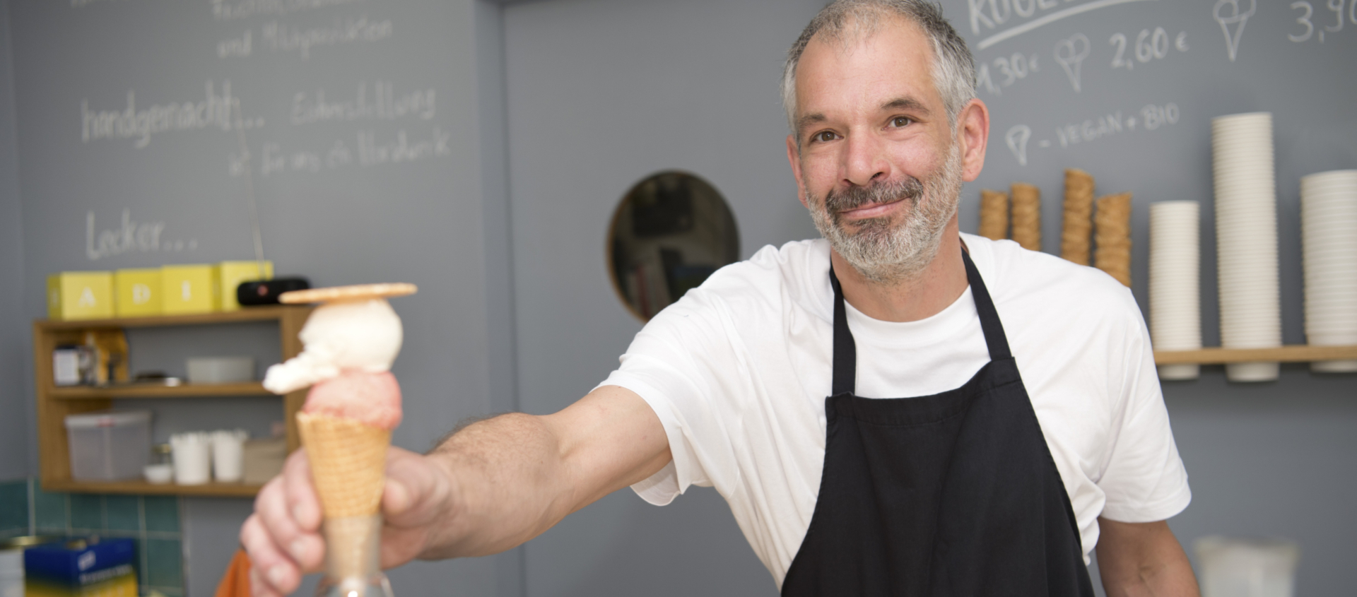 Eisverkäufer mit schwarzer Schürze reicht eine Eistüte mit zwei Kugeln über eine Glasvitrine hinweg