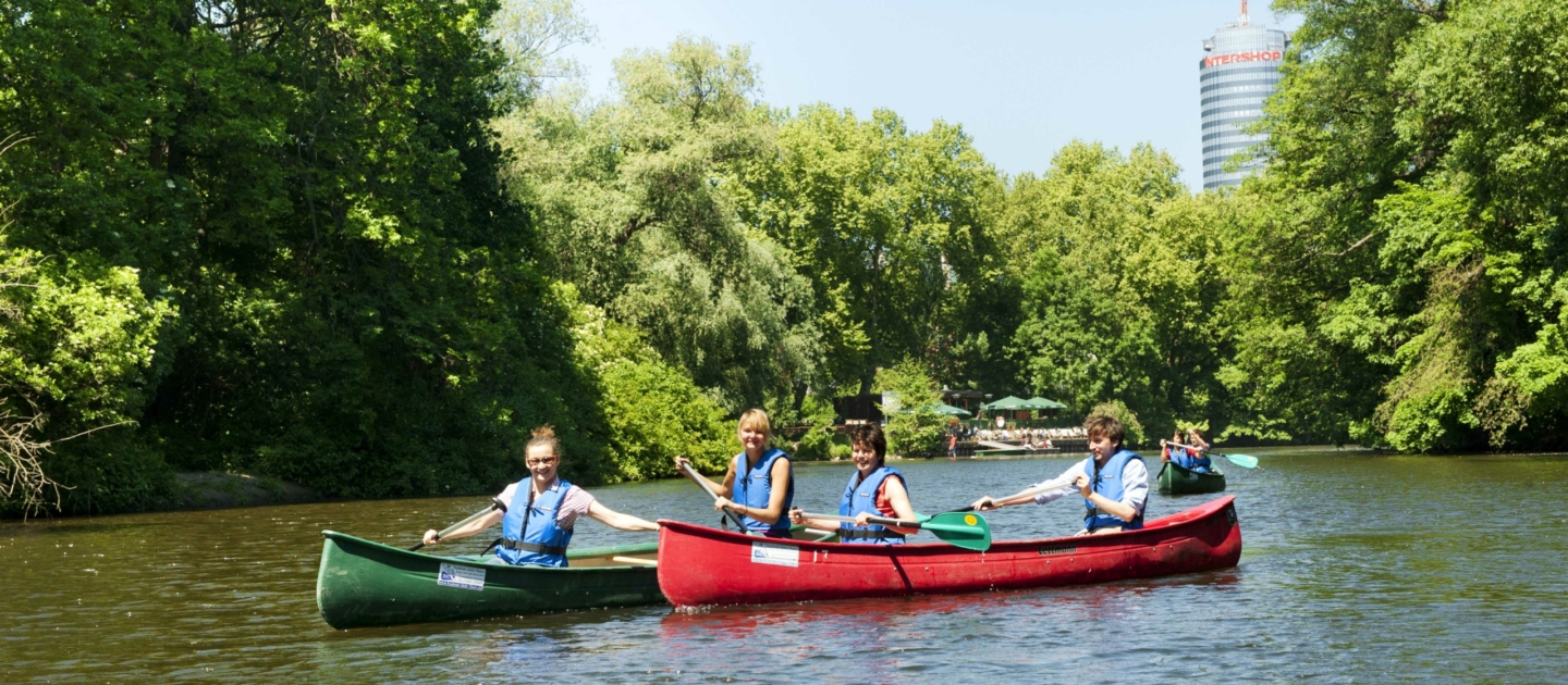 Zwei Kanus mit jeweils zwei Personen auf der Saale in Jena umgeben von grünen Bäumen und im Hintergrund der Jentower