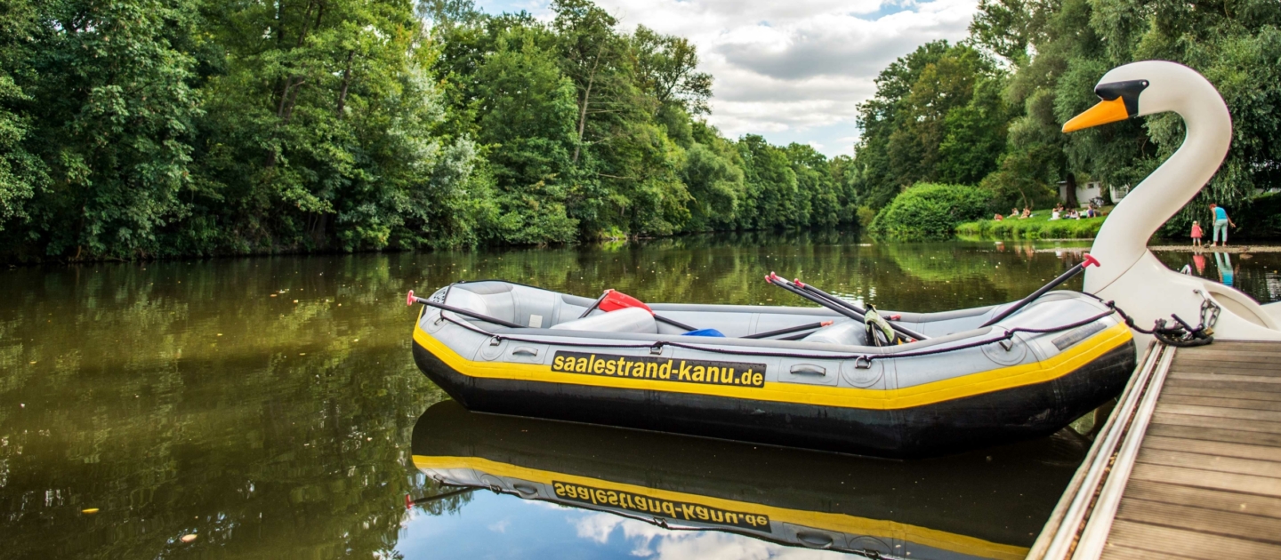 Schlauchboot mit der Aufschrift 'saalestrand-kanu.de' liegt an einem Steg an der Saale in Jena neben einem großen Schwan-Skulptur mit orangem Schnabel