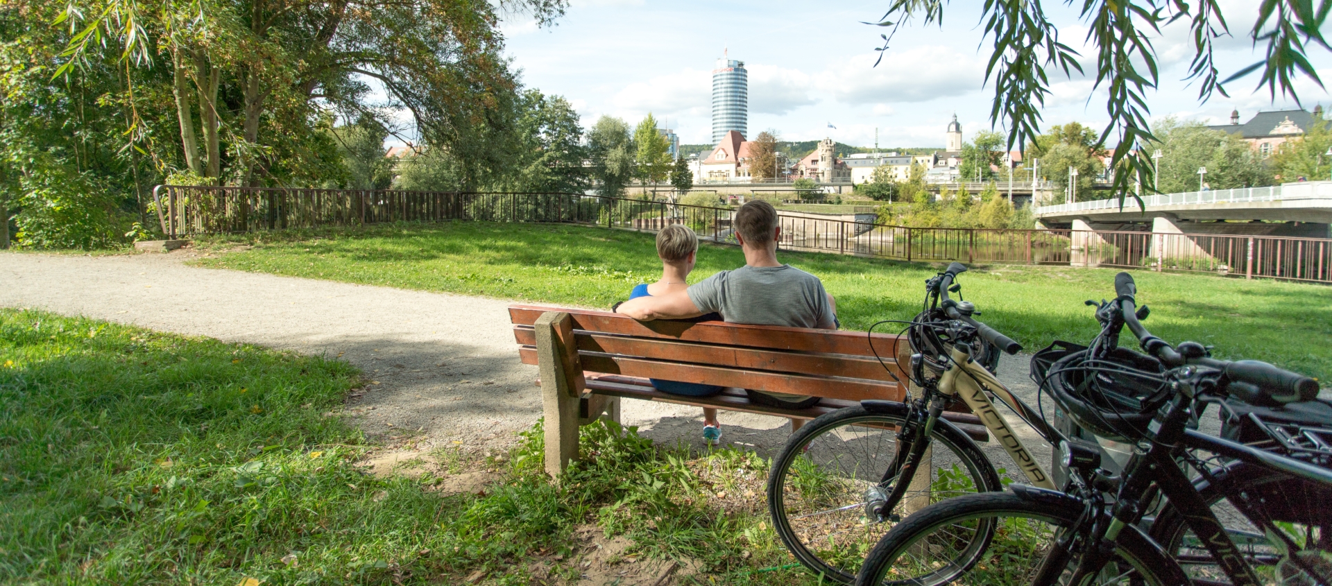 Zwei Personen sitzen auf einer Parkbank im Paradiespark mit Blick auf den Jentower, im Vordergrund stehen zwei Fahrräder auf einer Wiese