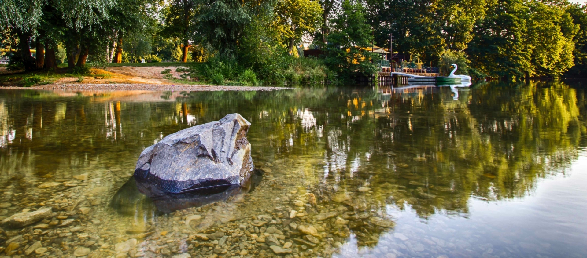 Flussufer der Saale mit klarem Wasser, einem großen Stein im Vordergrund und dichtem Baumbestand am Ufer, an dem ein Steg mit einem weißen Tretboot in Schwanform liegt
