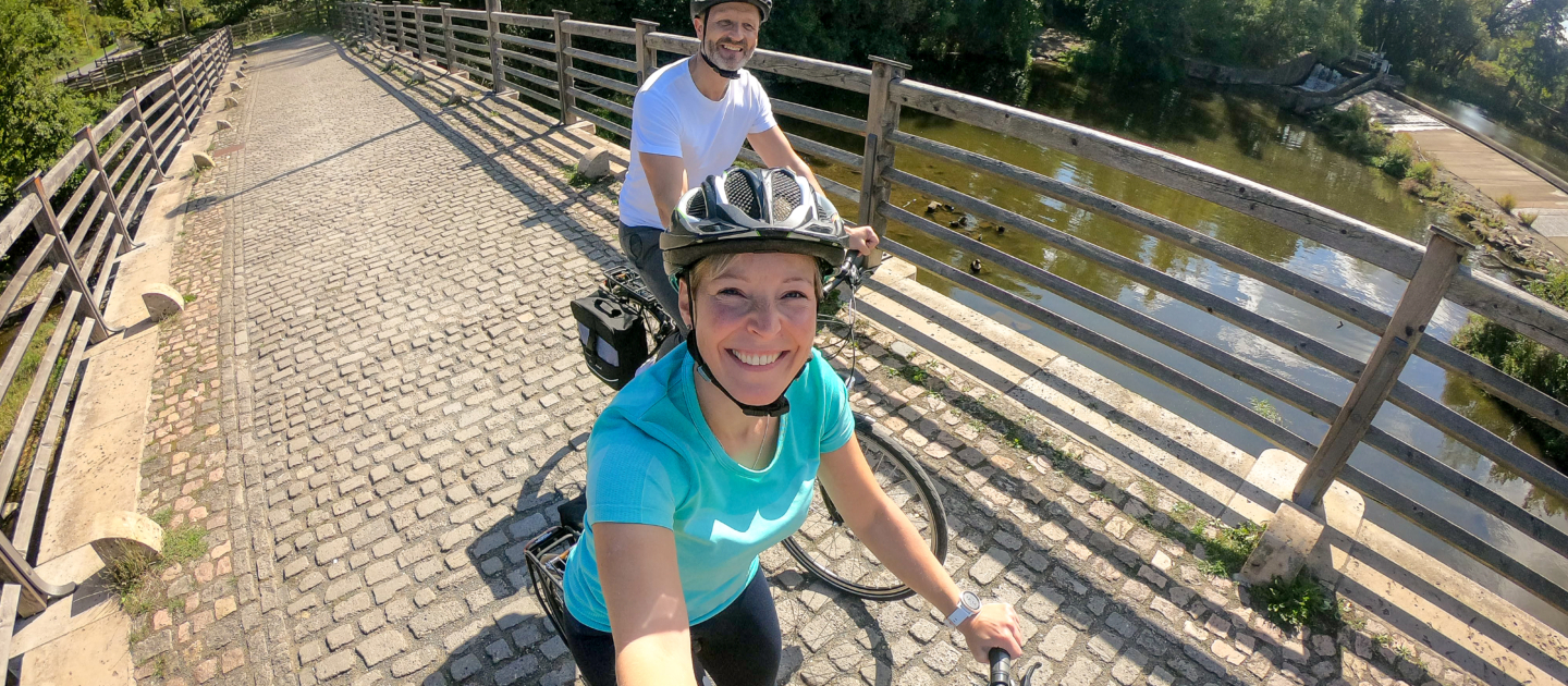 Zwei Radfahrer auf der gepflasterten Burgauer Brücke in Jena mit Holzgeländer und der Saale im Hintergrund