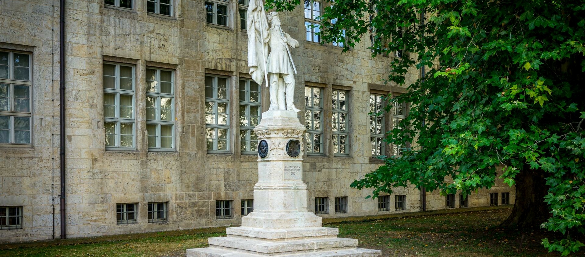 Das Burschenschaftsdenkmal vor der Universität Jena zeigt eine steinerne Statue eines stehenden Mannes mit erhobener Fahne auf einem mehrstufigen Sockel neben einem großen Baum.